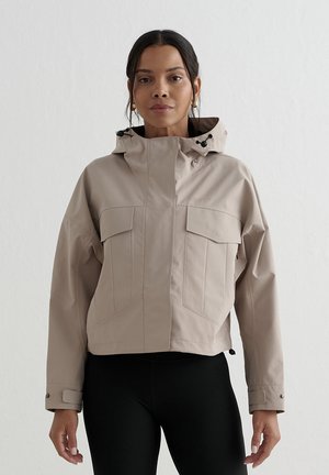 Femme portant une veste à capuche beige avec de grandes poches devant et un pantalon noir, debout devant un fond clair uni.