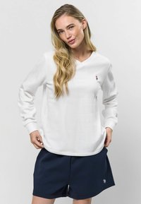 Biela mikina s dlhým rukávom a výstrihom do V s rebrovanými manžetami, s malým výšivkovým logom na hrudi, v kombinácii s námorníckymi šortkami.