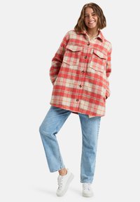 Rode en beige geblokte oversized flanellen shirt met knoopsluiting, twee borstzakken en een licht gestructureerde stof. Draagt als combinatie met lichtblauwe jeans.