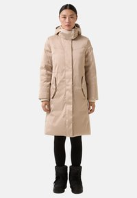 Beige gepolsterte Parka mit hohem Kragen, zwei seitlichen Taschen und einem Reißverschluss. Getragen über schwarzen Leggings und schwarzen Schneestiefeln.