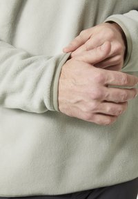 Fleece pullover in lichtgroen, met een zachte textuur en omgeslagen mouwen met een contrasterende binnenafwerking. Handen die de mouw vasthouden.