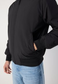 Black bomber jacket with a smooth texture, zip closure, ribbed cuffs, and side pockets. Paired with light blue jeans. Effective casual style.