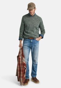 Grauer, gefleckter Pullover, blauer Jeanshemd, hellblaue Jeans, braune Schnürstiefel mit weißen Sohlen, hält ein rotes und beige kariertes Hemd.