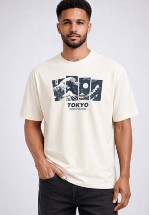Man with short curly hair and beard wearing a cream T-shirt with black graphic panels and "TOKYO" text, standing with one hand in pocket.