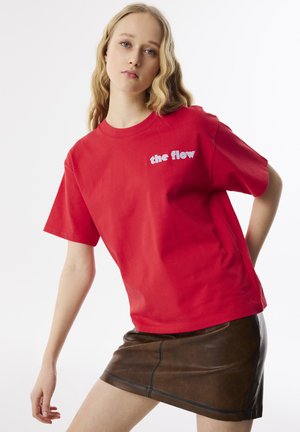 T-shirt en coton rouge à manches courtes avec le texte blanc "the flow" sur le côté gauche de la poitrine. Assortis à une mini jupe en faux cuir marron.