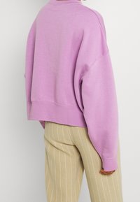 Person med en lös lavendel sweatshirt och beige byxor med vita vertikala ränder, står med ryggen mot kameran.