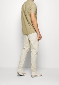 Camisa de manga corta en color caqui claro combinada con pantalones tapered en color crema y zapatillas altas blancas, mostrando un estilo limpio y casual.