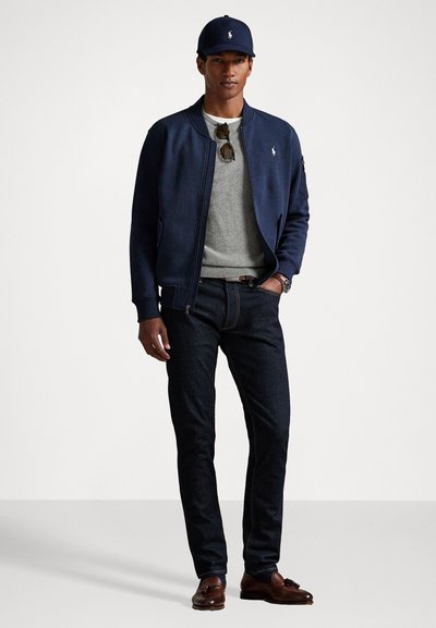 Navy bomwerjack over een lichtgrijze sweater, donkere spijkerbroek, bruine schoenen en een marineblauwe pet met een logo. Zonnebril op het shirt.