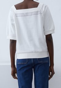 Vit tröja med fyrkantig halsringning och spetsdetaljer på baksidan. Kort puffärmar och ribbade manschetter. Matchas med mörk denimjeans.