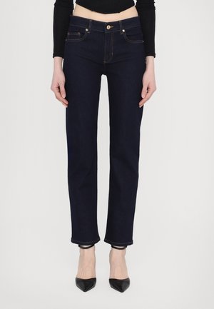 Jeans de mezclilla azul oscuro con un ajuste slim, cintura de altura media, cinco bolsillos, costuras claras y dobladillos sin rematar, combinados con zapatos de tacón negro en punta.