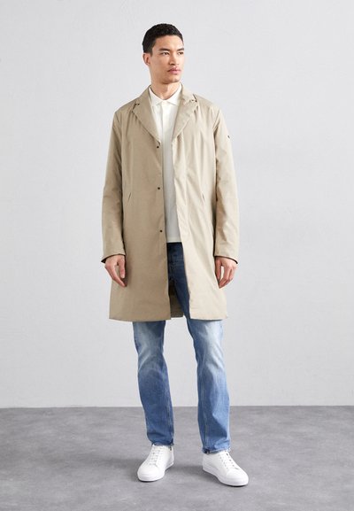 Beige, lightweight trench coat with a front button closure, side pockets, and a relaxed fit worn over a white polo and blue jeans. White sneakers.