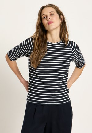 Woman with long brown hair wearing a black and white striped short-sleeve shirt and dark pants, standing with hands on hips.