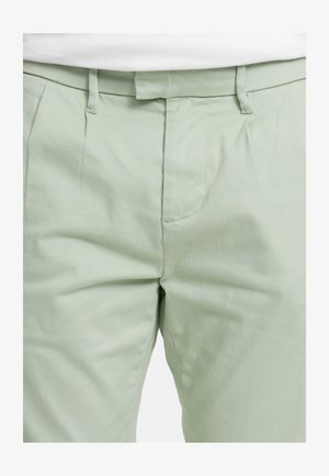 Světlé zelené kalhoty na míru s hladkou texturou. Mají přední záhyby a zapínání na knoflíky. Šití podél kapes pro detailní vzhled.