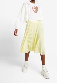 Chaqueta blanca corta, camiseta gráfica con diseño de reno y falda midi amarilla con plisados. Zapatillas gruesas blancas completan el conjunto.