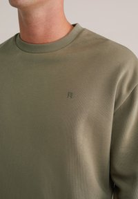 Olivno zelena trenirka z ribastim okroglim izrezom in majhnim vezenim logotipom. Gladka tekstura, sproščen kroj in dolgi rokavi.