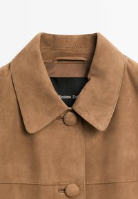 Braune Wildlederjacke mit breitem Kragen und zwei verdeckten Knöpfen, die ein teilweise sichtbares schwarzes Innenetikett zeigt.