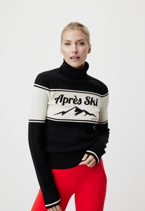 Femme blonde portant un pull à col roulé noir et blanc avec un motif "Après Ski" et montagne, associé à un pantalon rouge vif.