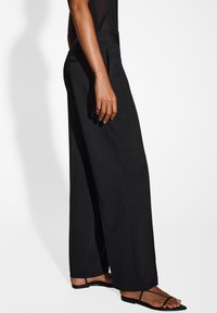 Pantalons larges noirs en tissu lisse avec une taille plate. Présente une taille cintrée et des fentes à l'ourlet. Portés avec des sandales minimalistes noires.