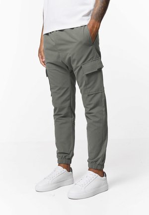 Mężczyzna w szarych joggerach cargo z elastycznymi mankietami i białych trampkach, stojący z jedną ręką w kieszeni na jednolitym białym tle.