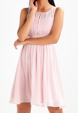 Kobieta w różowej, bezrękawowej sukience do kolan z plisami i marszczonym dekoltem, stojąca z jedną ręką skrzyżowaną, a drugą przy twarzy.