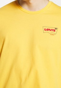 T-shirt di cotone giallo con scollatura rotonda. Presenta il logo Levi's in rosso, contornato in nero, nella zona del petto sinistro. Texture morbida.
