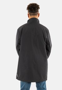 Sort vandtæt jakke med høj krave, lange ærmer og lige snit, båret over blå jeans. Ingen synlige mønstre eller hardwaredetaljer.