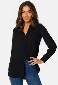 Blusa nera a maniche lunghe con colletto, tessuto morbido, polsini con bottone e orlo posteriore leggermente allungato, abbinata a jeans blu.