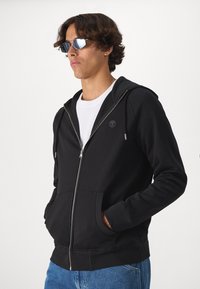 Felpa nera con zip, logo piccolo, tasche laterali, cappuccio regolabile con cordino e polsini a costine, abbinata a jeans blu. Tessuto della felpa liscio.