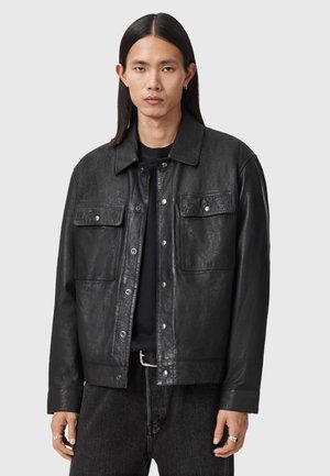 Man met lang donker haar draagt een zwarte leren jas over een zwart shirt en donkere jeans, staand tegen een effen lichte achtergrond.