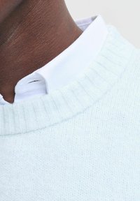 Lys blå strikket sweater med ribbet halslinning, lagt over en hvid skjorte med krave. Glat tekstur med detaljeret syning.