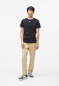 Černé tričko s krátkým rukávem a bílým lemováním límce, v kombinaci s béžovými chinos kalhotami a námořnickými teniskami s bílými detaily.