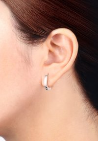 Silberner Creolenohrring mit einer glatten, polierten Oberfläche. Verfügt über ein elegantes, gewelltes Design und einen minimalistischen Stil, der eng am Ohrläppchen sitzt.