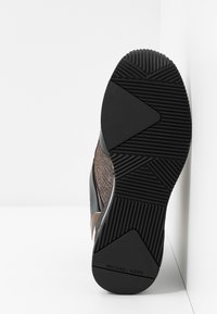 Semelle de chaussure avec une accroche en caoutchouc noir texturé, motifs géométriques de la bande de roulement et le nom de la marque "Michael Kors" près du talon.