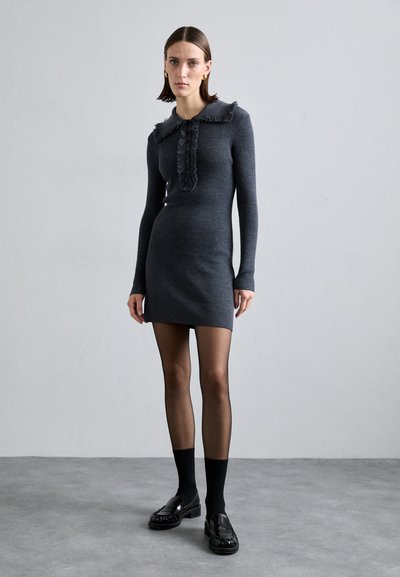 Gray ribbed knit dress with a ruffled collar and front placket. Model wears black tights and loafers with a glossy finish. Simple design.