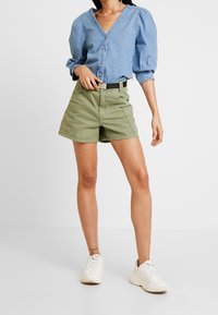Camicetta di jeans azzurra chiara con maniche a sbuffo, chiusura con bottoni, abbinata a pantaloni corti verde oliva con tasche frontali e una cintura nera. Sneakers bianche.