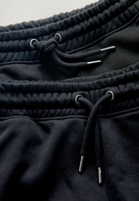 Pantaloni della tuta neri con vita elastica, coulisse con punte in metallo, caratterizzati da una texture liscia e vita a coste. Due paia distesi in piano.