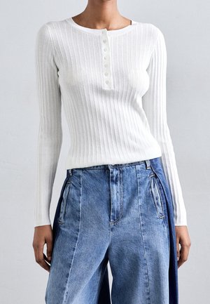 Personne portant un haut blanc côtelé à manches longues boutonné, rentré dans un pantalon en denim bleu taille haute à jambes larges avec des poches avant.