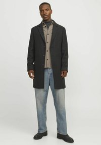 Cappotto nero in lana con texture e collo a ve, abbinato a una camicia grigia con bottoni e jeans a gamba larga azzurri, indossato con scarpe nere.