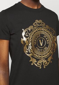 Černé tričko s velkým zlatým fóliovým kruhovým logem s ornamentálními detaily. Design obsahuje text "VERSACE JEANS COUTURE" a centrální "V".