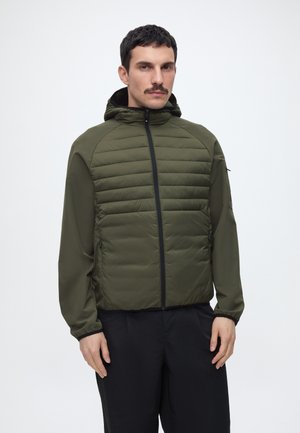 Mann mit dunklen Haaren und Schnurrbart, der eine olivgrüne Steppjacke mit Kapuze und schwarze Hose trägt, steht vor einem einfarbigen Hintergrund.