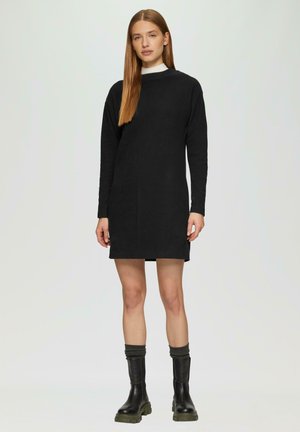 Schwarzes langärmliges Kleid mit rundem Halsausschnitt, das eine gerade Silhouette und glattes Material aufweist, kombiniert mit knielangen schwarzen Stiefeln und grauen Socken.