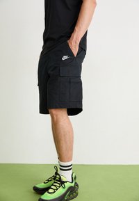 Černé cargo šortky s kapsami, s bílým logem Nike, doplněné zeleno-černými teniskami a proužkovanými ponožkami. Hladká textura, délka po kolena.