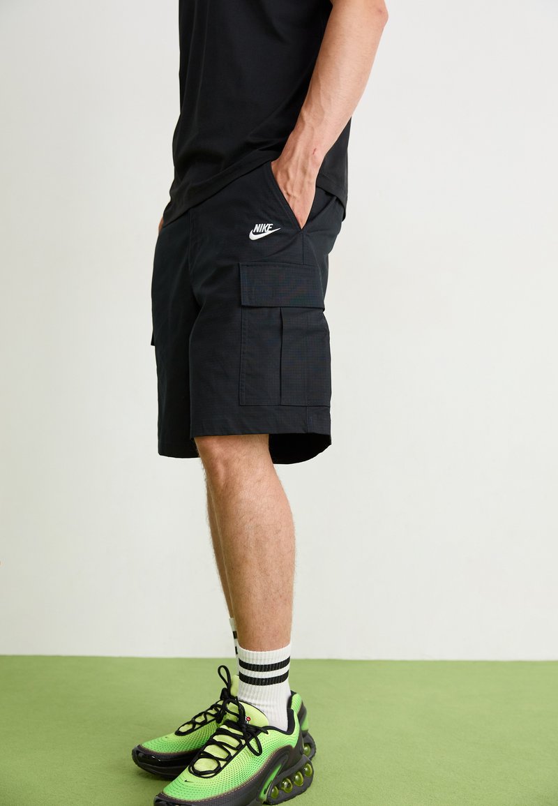 Bermudas negras con bolsillos, con un logo blanco de Nike, combinadas con zapatillas verde y negras y calcetines de rayas. Textura plana, hasta la rodilla.