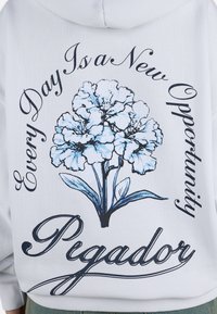 Hanorac alb cu un design floral albastru și text scris. Graficul include fraza „Fiecare zi este o nouă oportunitate” și „Pigador.”