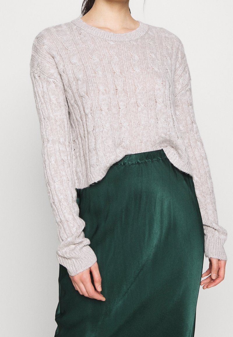 Femme portant un pull torsadé gris clair et une jupe en satin vert foncé, montrée du cou jusqu’aux cuisses sur un fond uni.