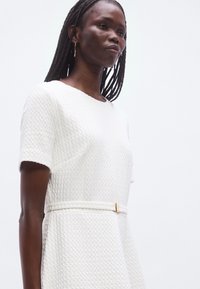 Robe blanche texturée avec manches courtes, taille cintrée et détails en métal doré. Le tissu présente un motif tissé subtil.