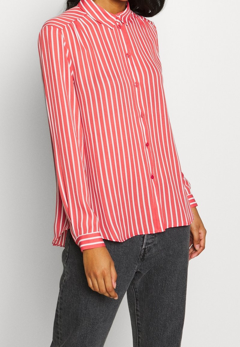 Red and white striped long-sleeve shirt with a collared neck, front buttons, and cuffed sleeves, paired with dark jeans.