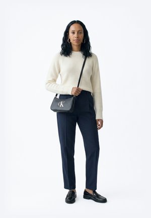 Nő áll, krémszínű bordázott pulóvert, tengerészkék szabott nadrágot, fekete mokaszint és ezüst CK logós fekete keresztpántos táskát visel.