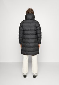 Svart puffjacka med hög krage, vertikalt quiltad design, knälång fåll och ribbade muddar. Buren över ljusa byxor och skor.