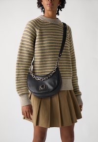 Borsa a tracolla in pelle nera con forma curvata, accento in catena argentata e dettaglio del logo. Indossata sopra un maglione a righe e una gonna beige.